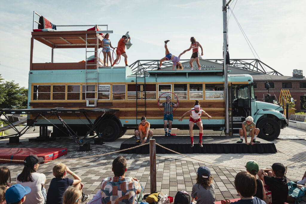 Acrobats jump and flip on top of and next to a turquoise school bus with a wooden center.