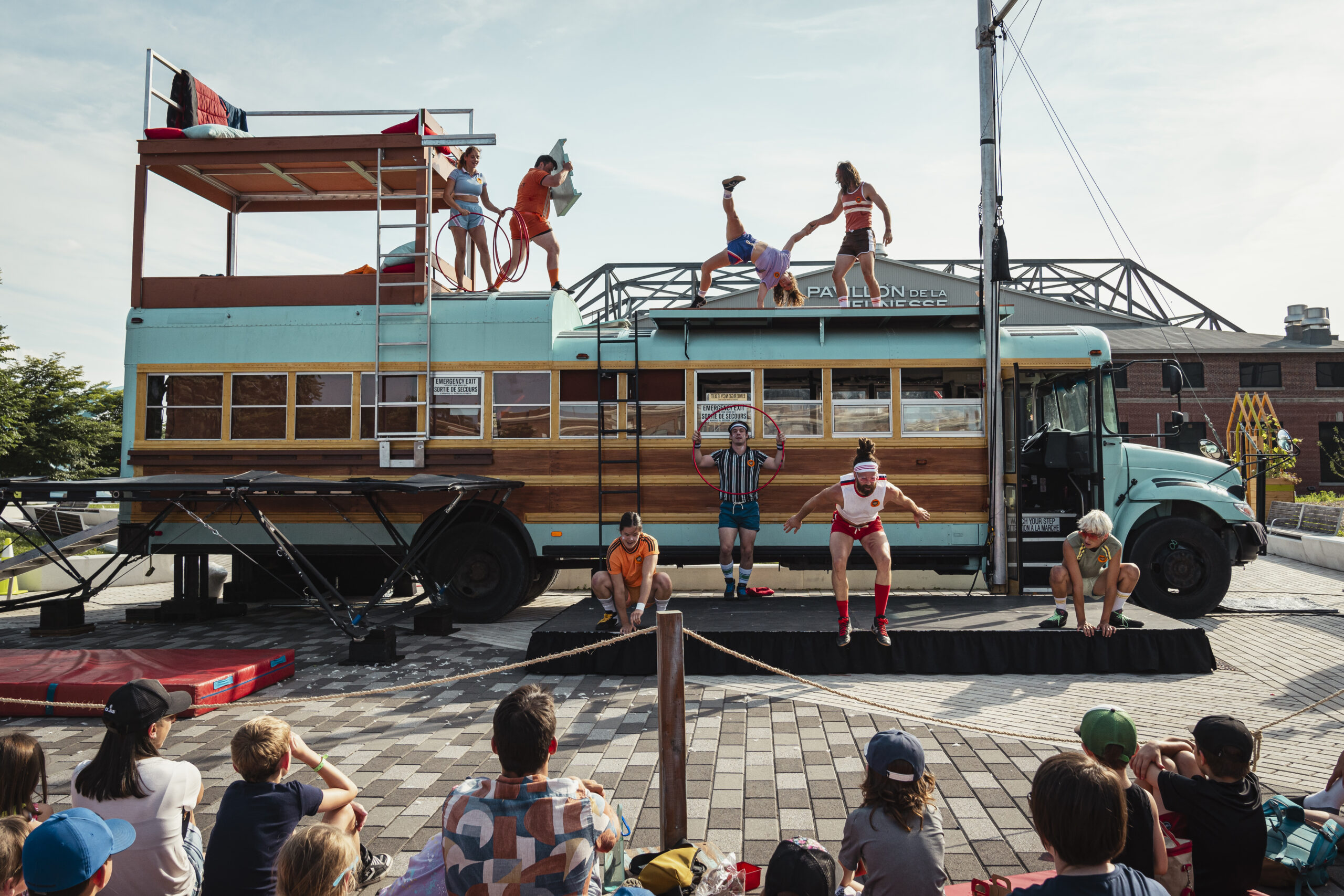 Acrobats jump and flip on top of and next to a turquoise school bus with a wooden center.