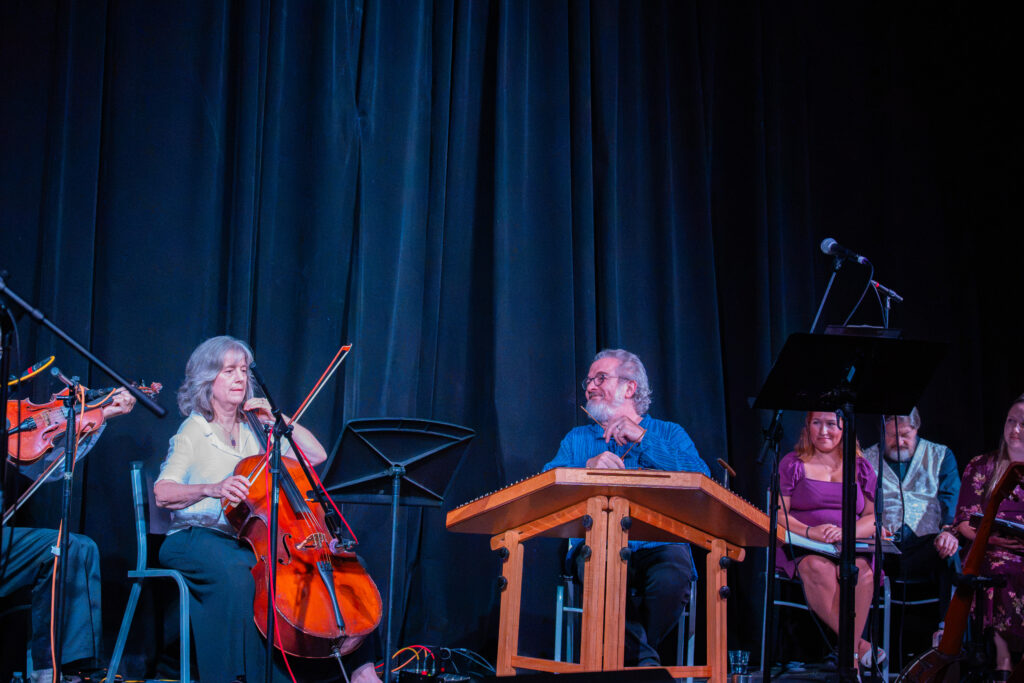 Glenn McClure sits in the center of the photo at a hammered dulcimer, holding the mallets and smiling at the musicians to his right side. The choir sits quietly on the other side.