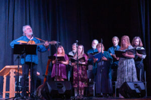 Composer Glenn McClure plays the guitar on the left, and there is a small choir on the right side.